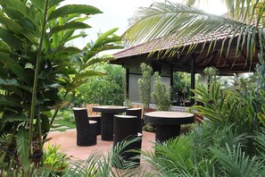An outdoor seating area with round tables and chairs, surrounded by lush greenery and a traditional-style building with a tiled roof. - Abhishta Agri Farm Stay (Kunigal)