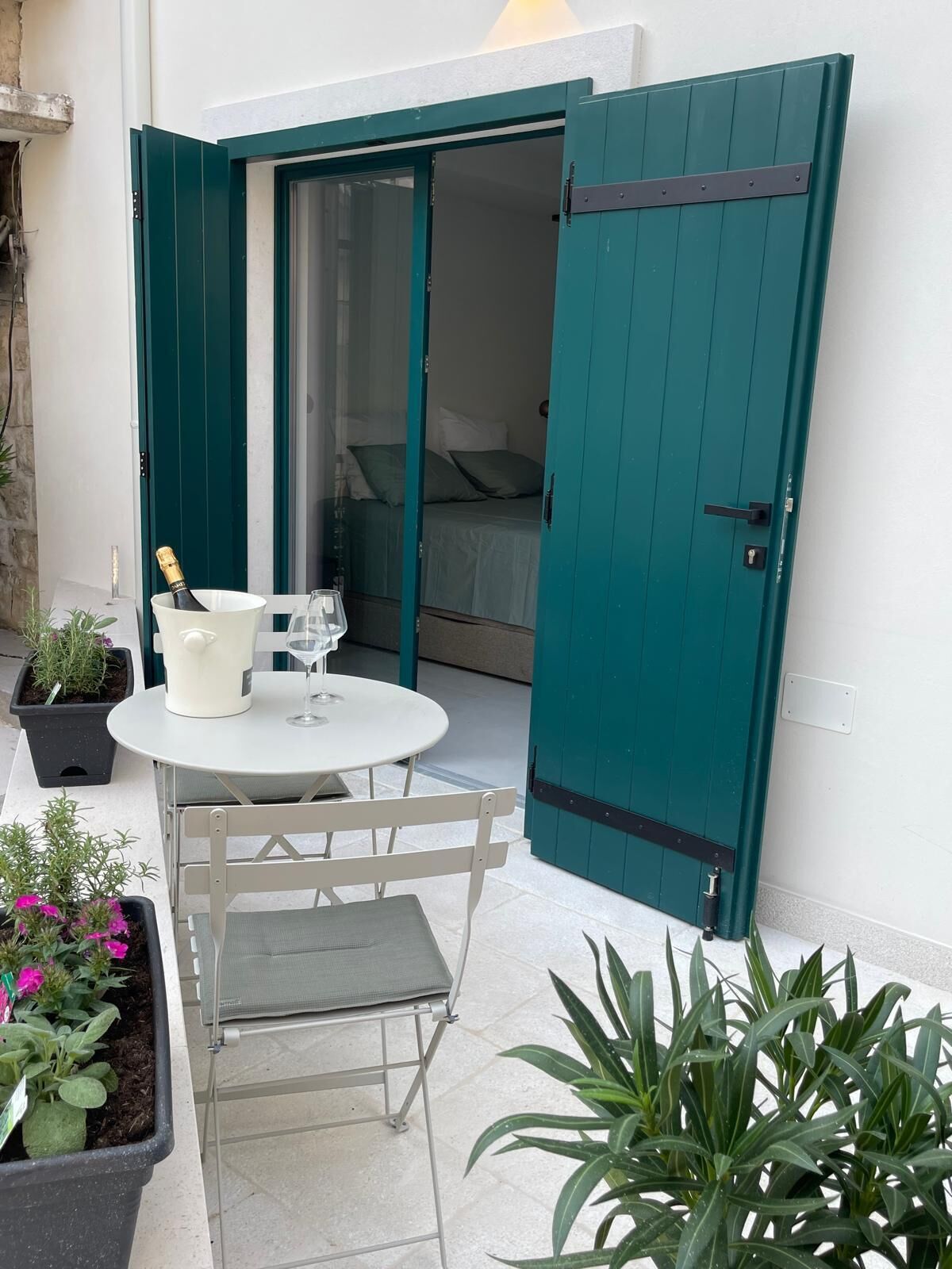 A small outdoor seating area with a table and chairs, a bottle of champagne, and potted plants.