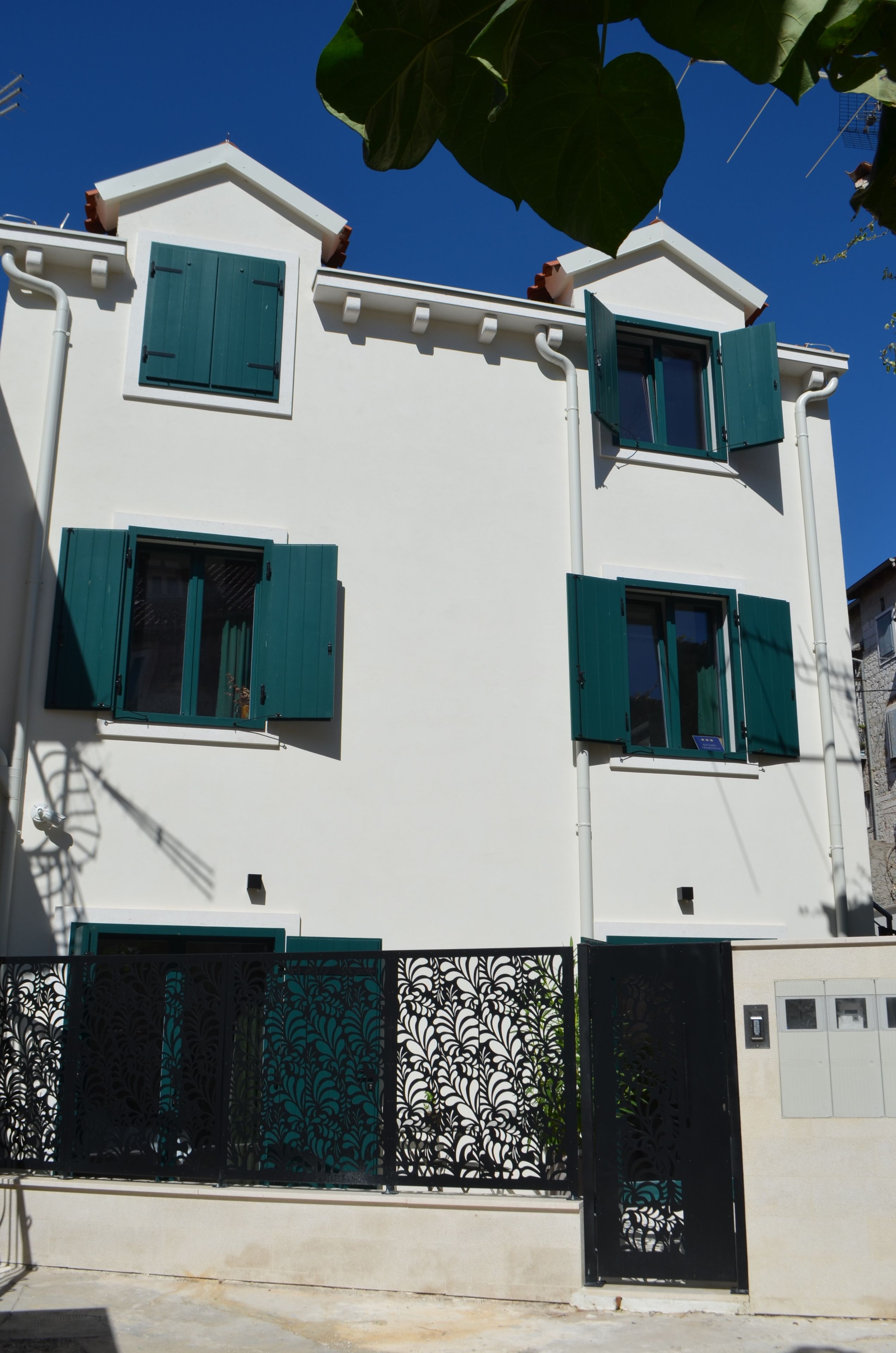A two-story building with green shutters, a white facade, and a decorative metal gate.