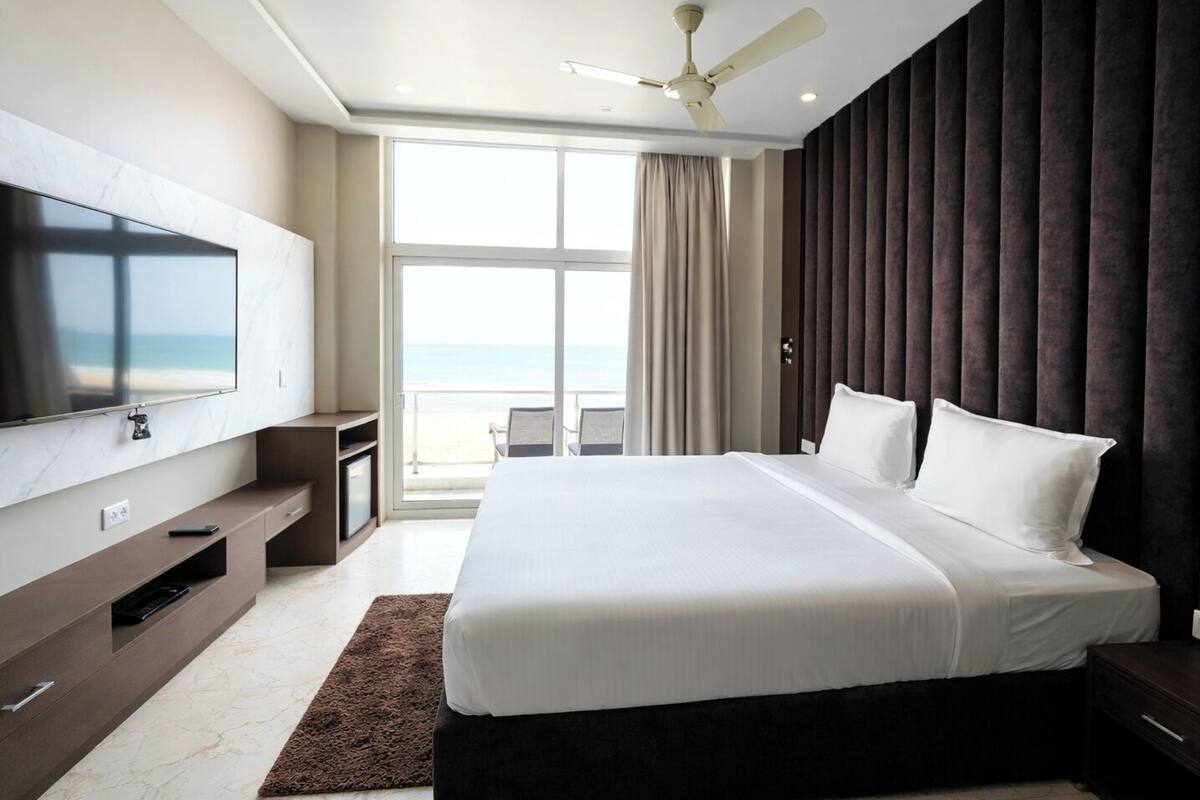 A modern hotel room with a large bed, a flat-screen TV, a ceiling fan, and a view of the ocean.