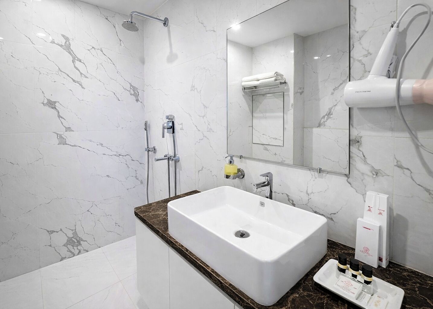 A modern bathroom with a marble wall, a white sink, a handheld showerhead, a towel rack, and a hairdryer.