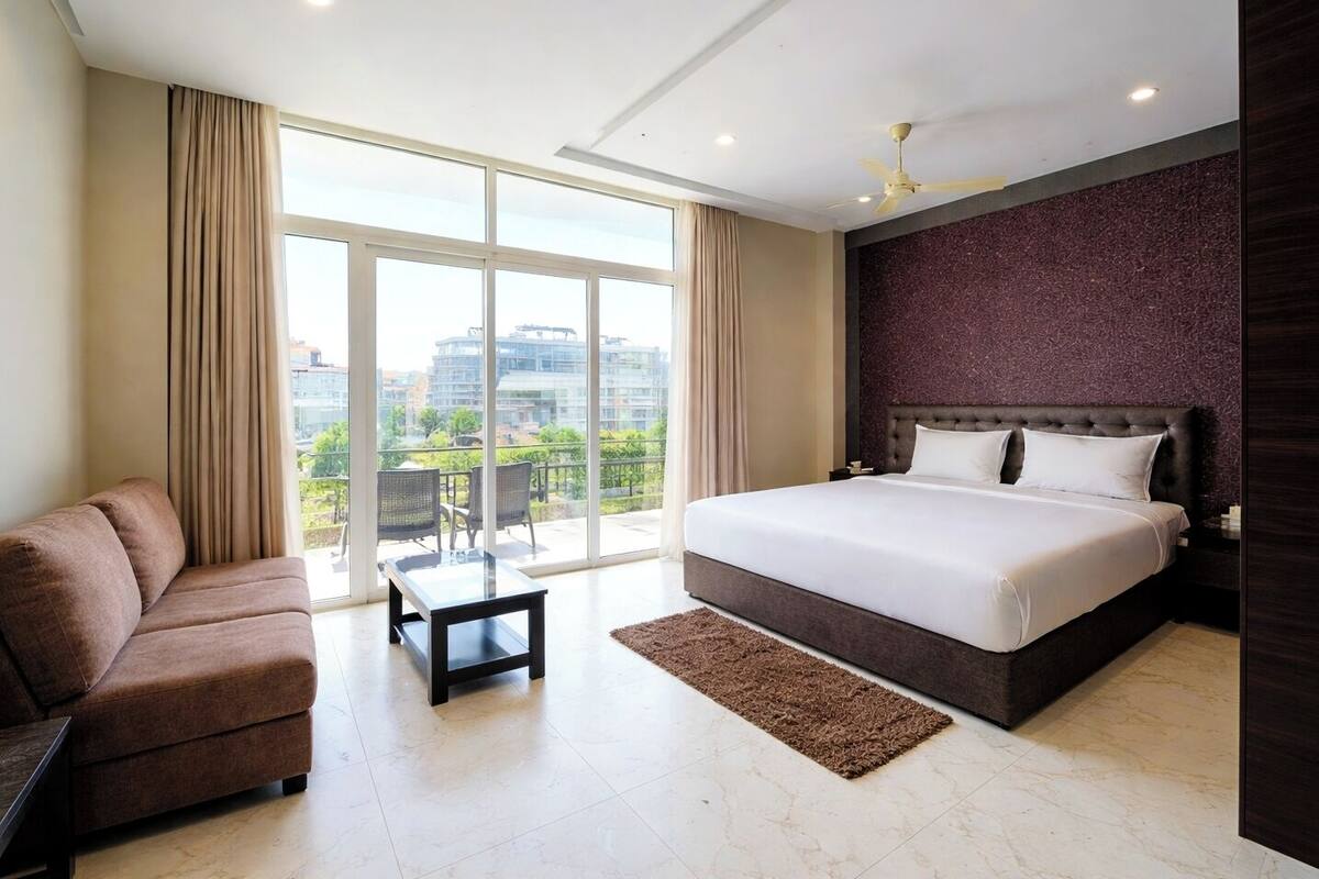 A modern hotel room with a large bed, a sofa, a small table, and a view of the cityscape through large windows.