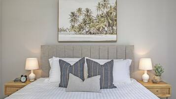 A neatly made bed with striped bedding, two bedside tables with lamps, a framed picture of a beach scene, and a small plant on one table.
