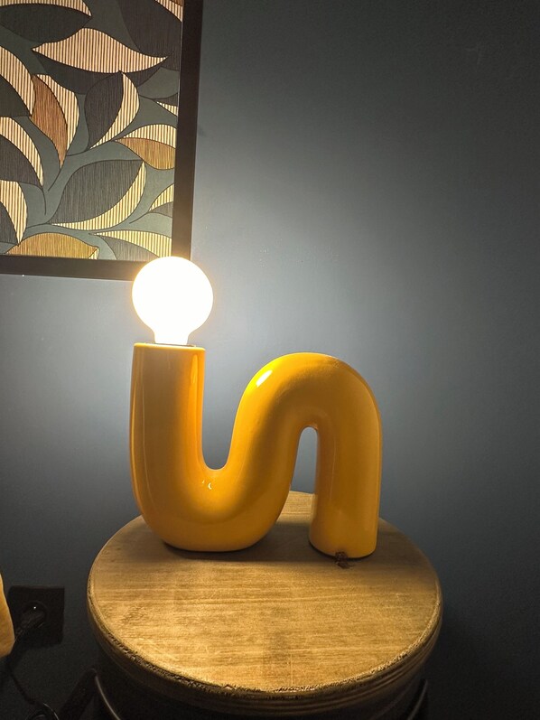 A yellow, curved lamp on a wooden table against a dark wall with a leaf-patterned artwork. - Le Loft des toits de Vernet (Vernet-les-Bains)