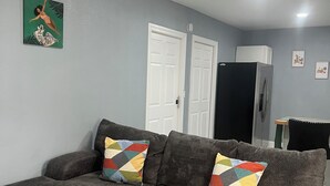 A grey sectional sofa with colorful pillows, a white door, a refrigerator, and a wall-mounted clock. - Cozy Haven Apartment (Winter Haven)