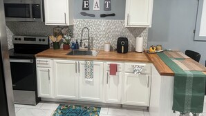 A modern kitchen with white cabinets, a stainless steel microwave, and a sink. There are decorative letters on the wall and a wooden countertop. - Cozy Haven Apartment (Winter Haven)