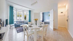 Ett modernt vardagsrum med en soffa, ett matbord och stolar. Det finns ett fönster med gardiner, en vas med blommor och en dörr som leder till ett annat rum.