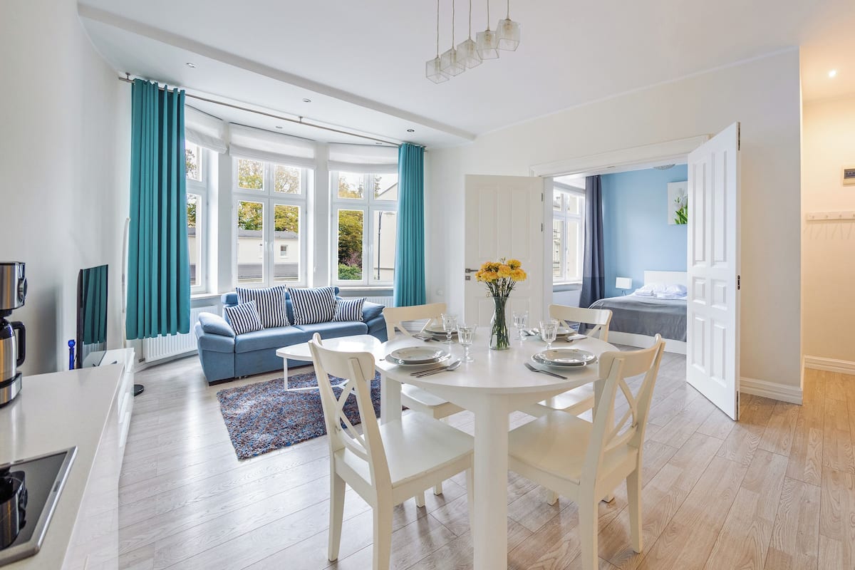 Una sala moderna con sofá, mesa de comedor y sillas. Hay una ventana con cortinas, un jarrón con flores y una puerta que da a otra habitación.