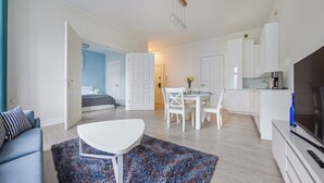 En modern lägenhet med vardagsrum, matplats och kök. I bakgrunden syns en säng, ett vitt matbord med stolar och ett kök med vita skåp och vitvaror.