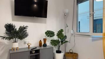 A modern living room with a flat-screen TV mounted on the wall, a grey entertainment unit, and two large potted plants.