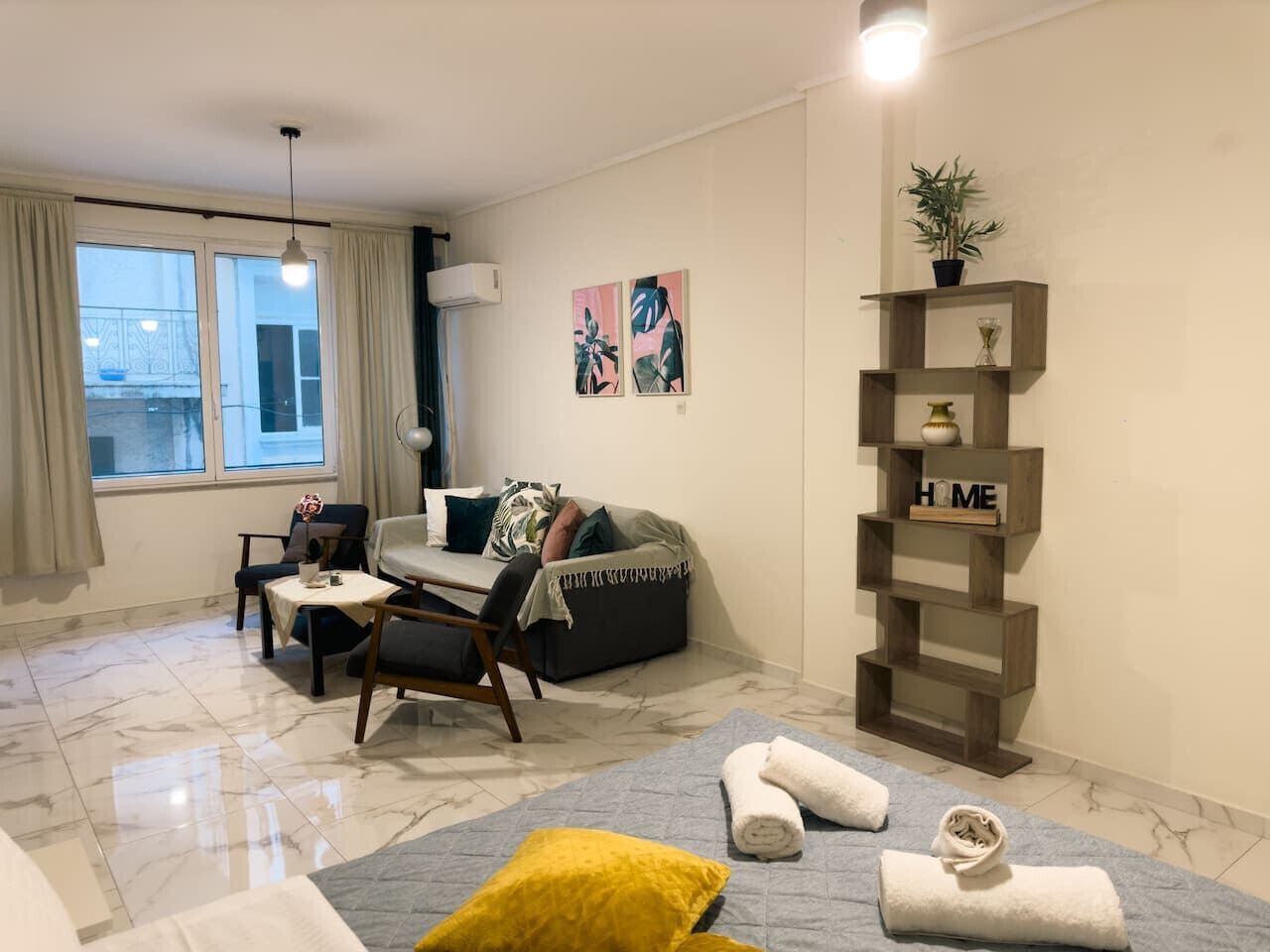A modern living room with a sofa, armchair, and coffee table. There is a window with curtains, a plant on a shelf, and artwork on the wall.