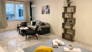 A modern living room with a sofa, armchair, and coffee table. There is a window with curtains, a plant on a shelf, and artwork on the wall. - Nikitara Nook-Close to Metro and Train (Athina)