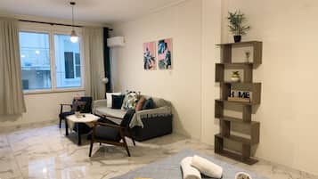 A modern living room with a sofa, armchair, and coffee table. There is a window with curtains, a plant on a shelf, and artwork on the wall.