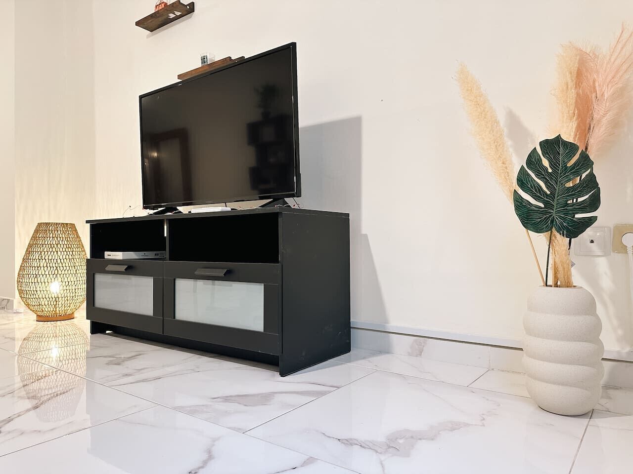 Un salon moderne avec un meuble de rangement noir pour les appareils électroniques, un grand téléviseur à écran plat, un lampadaire au design original et une plante décorative dans un vase blanc.