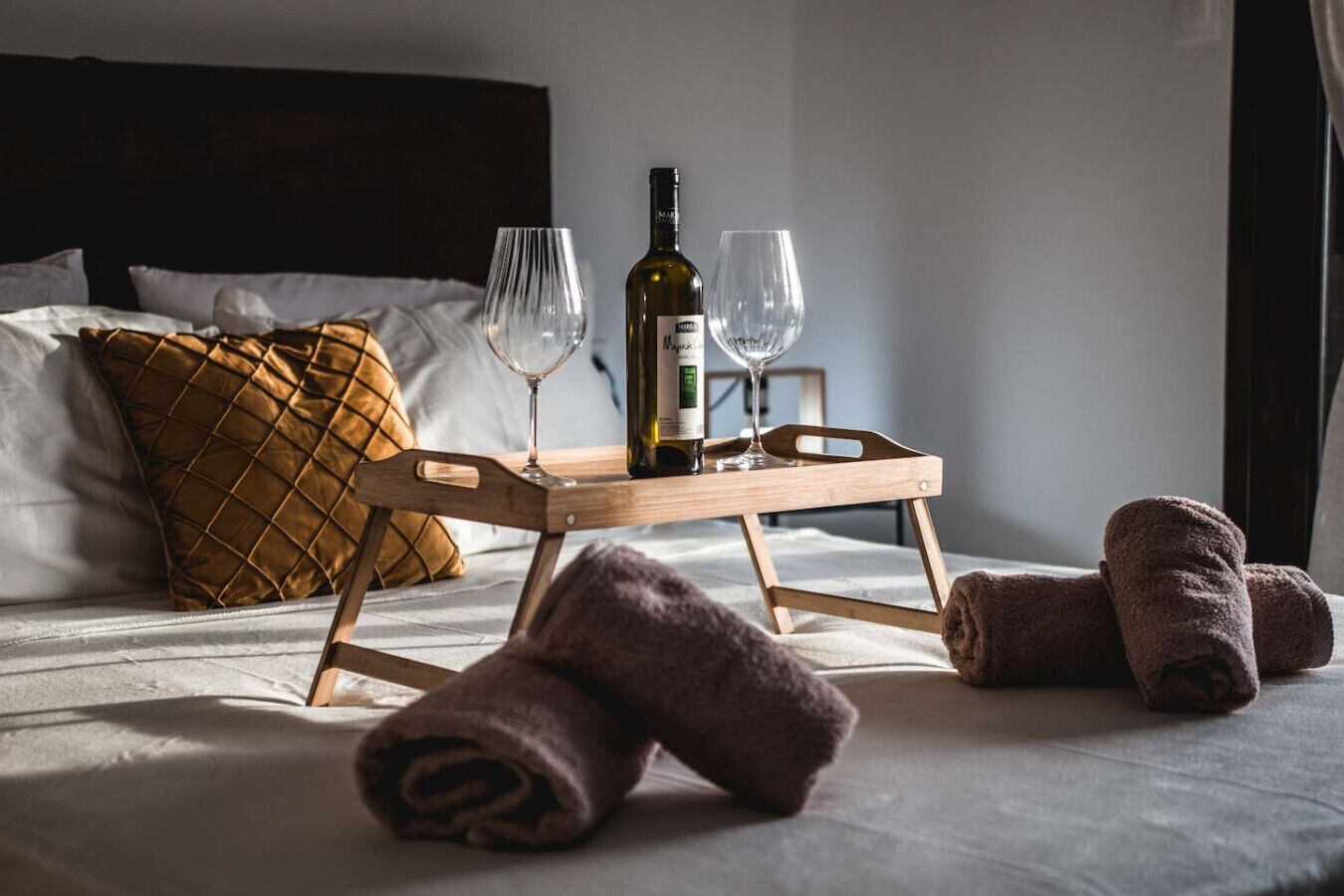 Ein ordentlich gemachtes Bett mit einem hölzernen Tablett, das eine Flasche und zwei Gläser trägt, ein braunes gestepptes Kopfkissen und aufgerollte Handtücher.