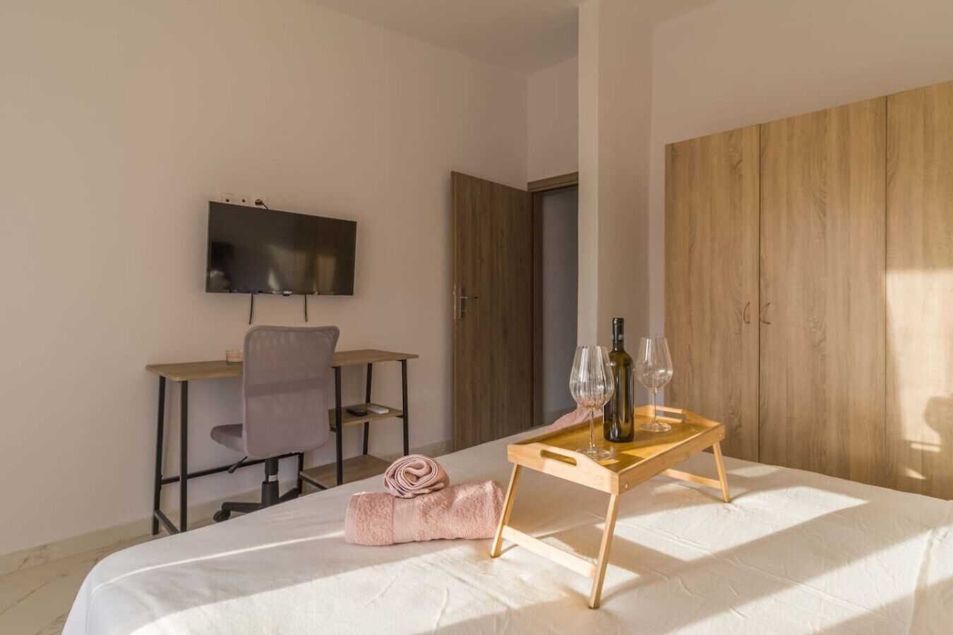 A bedroom with a bed, a desk, a chair, a TV, and a tray with glasses and a bottle.