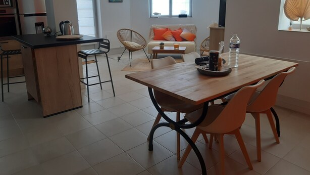 Eine moderne Küche mit Kochinsel, ein Essbereich mit Holztisch und orangefarbenen Stühlen sowie eine Wohnbereich mit Sofa und Kissen.