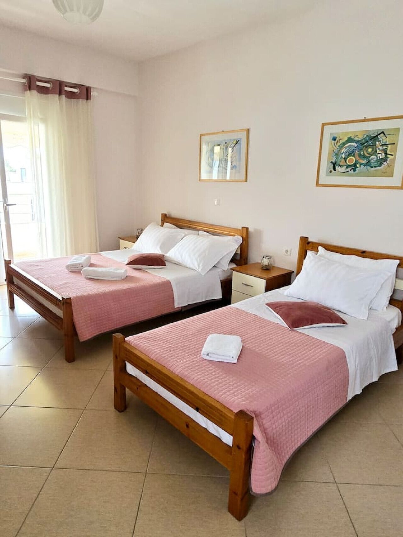 Habitación de hotel con dos camas de madera, cada una con colcha acolchada rosa y ropa de cama blanca. Hay dos mesitas de noche con lámparas, una ventana con cortinas y dos obras de arte enmarcadas en la pared.