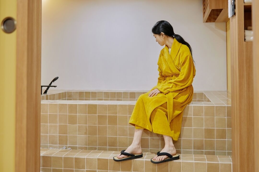 A person sitting on a tiled bench in a room with a wooden door frame and a tiled floor.