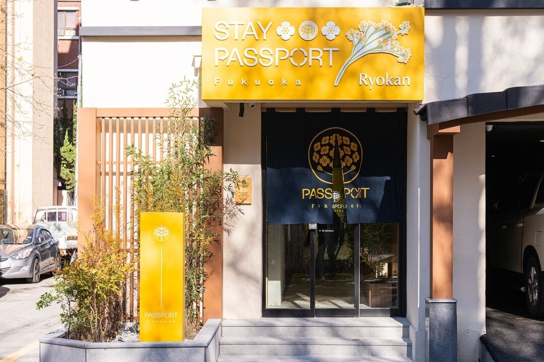 A hotel entrance with a yellow sign reading "STAY PASSPORT Fukuoka Ryokan".
