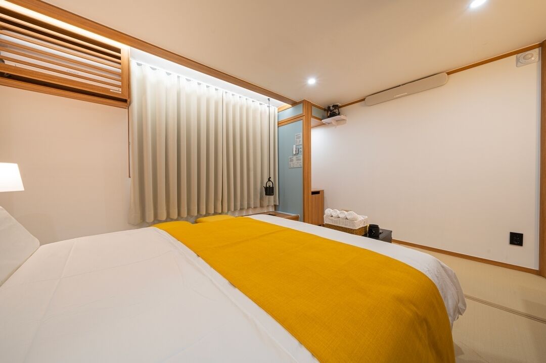 Habitación de hotel con cama, una manta amarilla, ventana con persianas, mesita de noche y una pequeña zona de estar con una silla y una mesa.