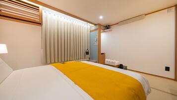 Habitación de hotel con cama, una manta amarilla, ventana con persianas, mesita de noche y una pequeña zona de estar con una silla y una mesa.
