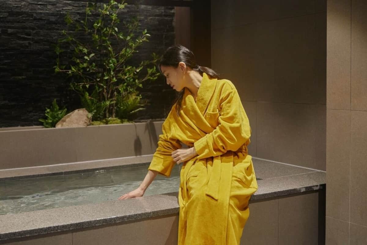 A person in a yellow robe standing by a water feature with a stone wall and greenery in the background.