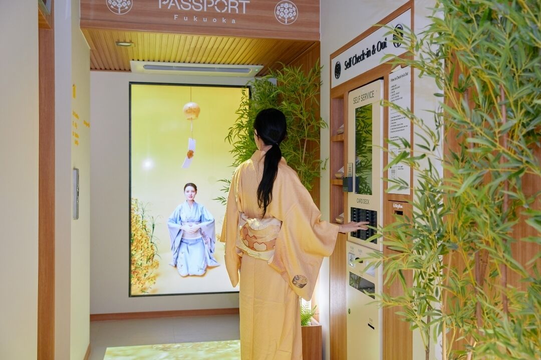 Una mujer con atuendo tradicional en una oficina de pasaportes, con un quiosco de autoservicio y un cartel de una mujer con un kimono azul.