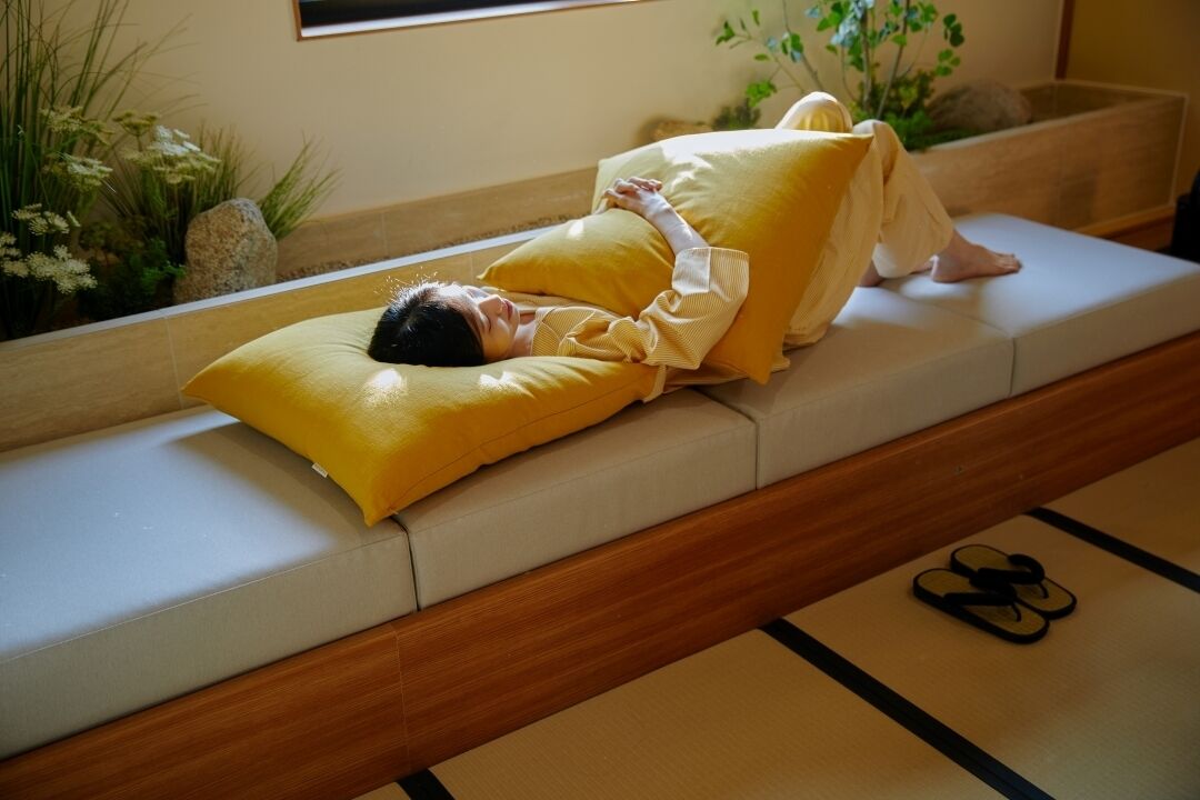 Una persona recostada en un sofá con almohadas amarillas, un cabecero de madera y un par de chanclas en el suelo.