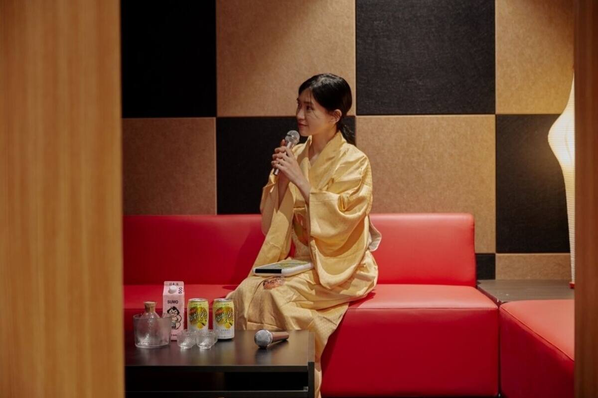 A person in a yellow robe sitting on a red couch with a microphone, holding a book, and a table with drinks and a box in front of them.