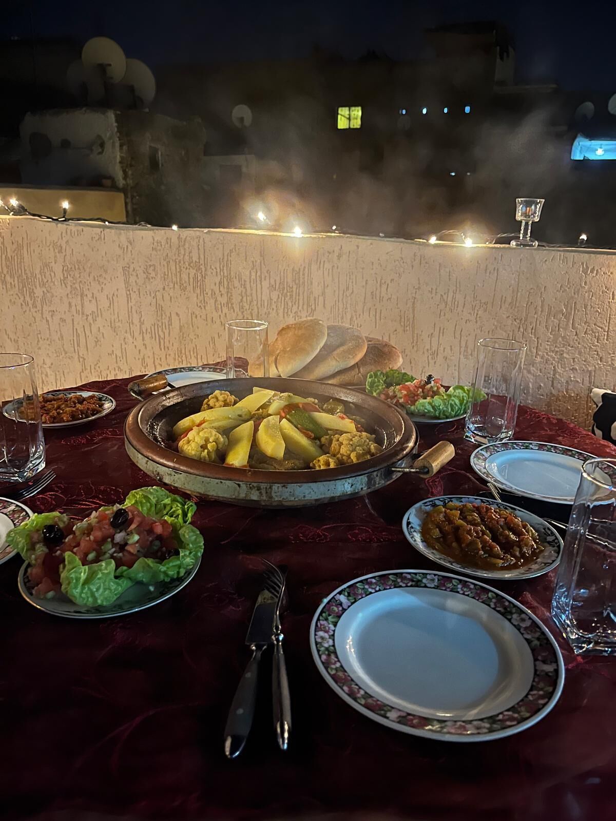 A dining table set with various dishes, including a large pot of stew, bread, and salads, on a terrace with a view of buildings and string lights in the background.