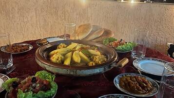 A dining table set with various dishes, including a large pot of stew, bread, and salads, on a terrace with a view of buildings and string lights in the background.