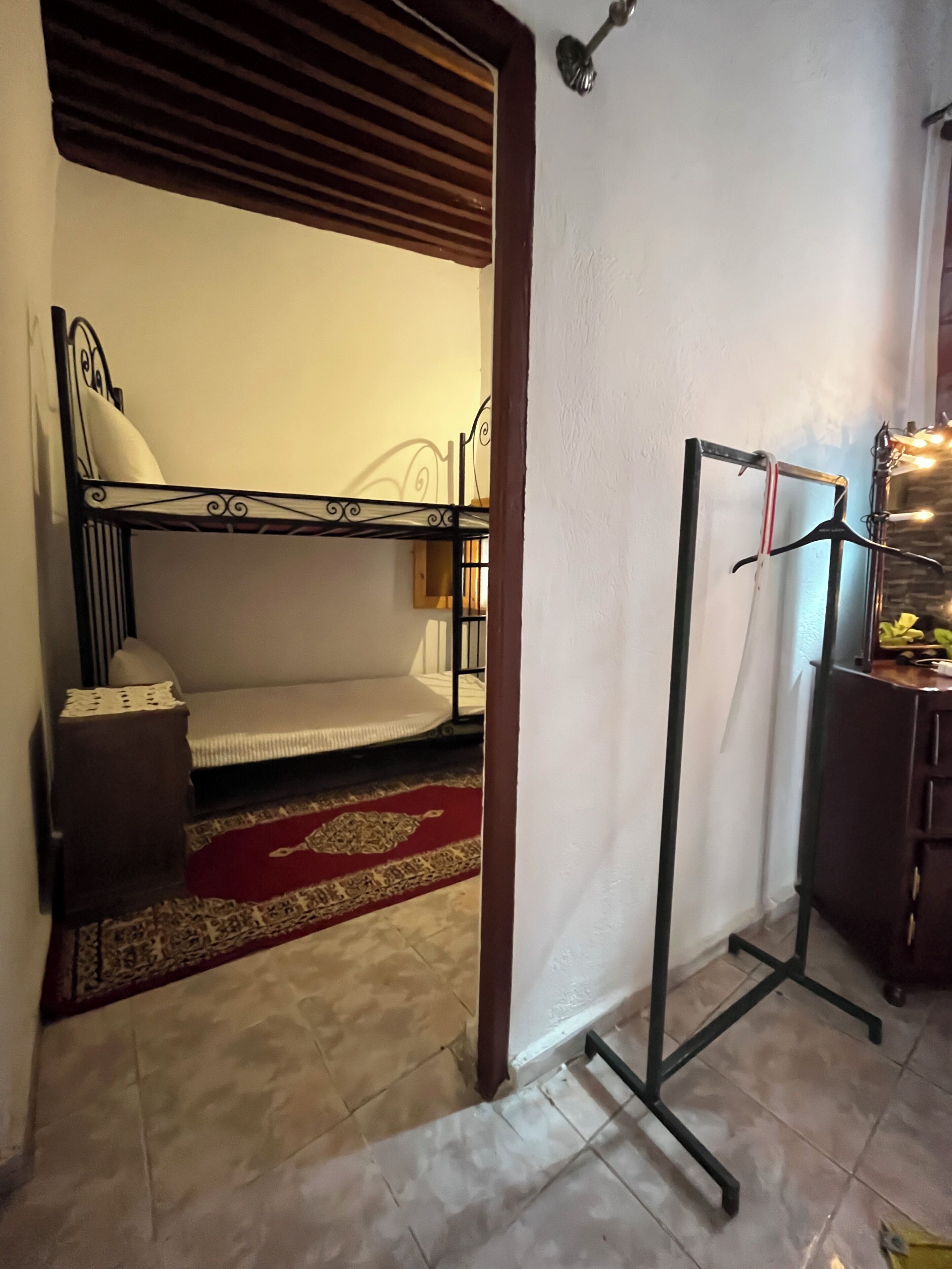 A compact room with a bunk bed, a small table with a lamp, a red patterned rug, and a metal rack with clothes.