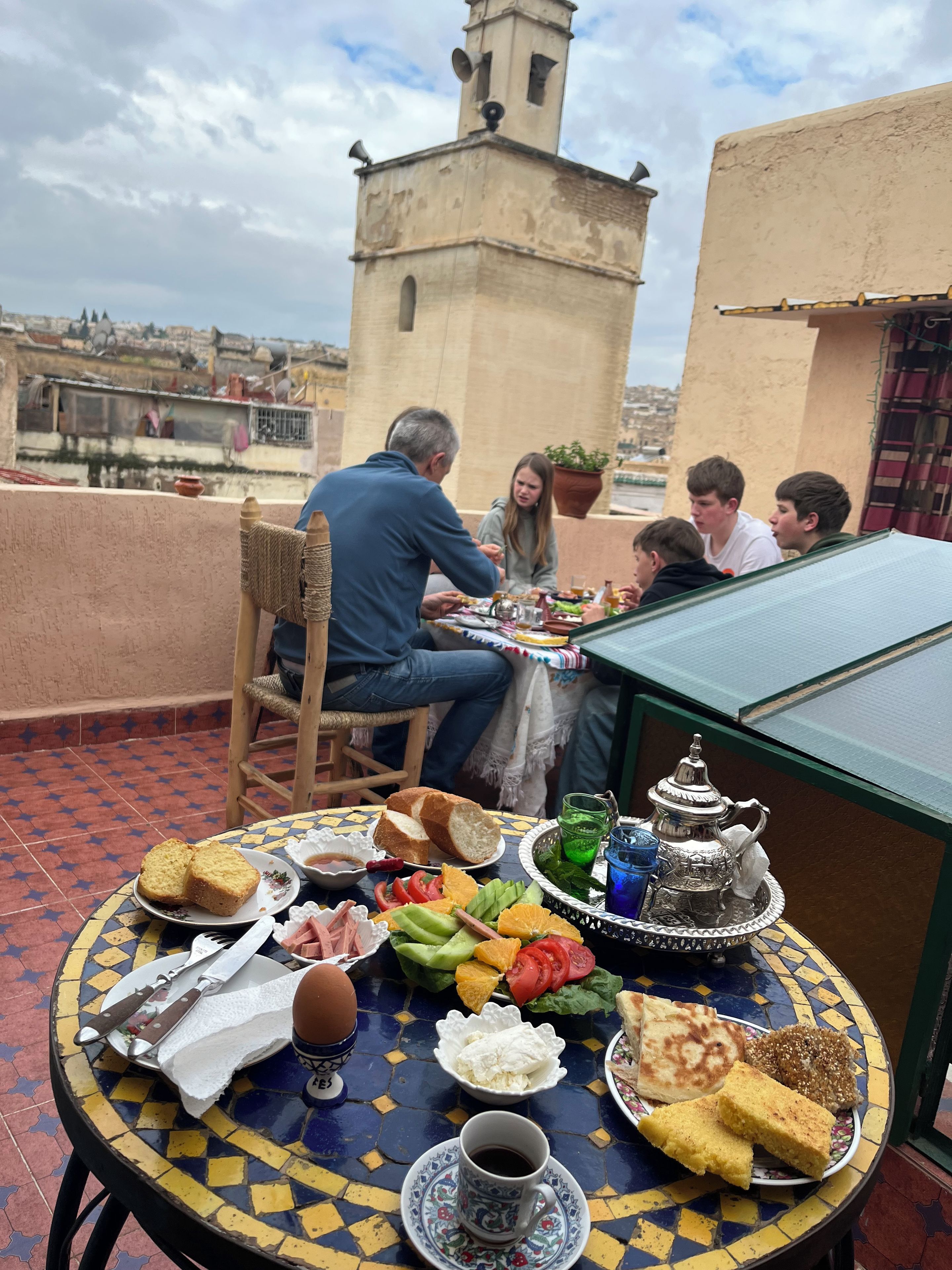 A group of people dining at a table with a variety of foods and drinks, including bread, vegetables, and a teapot, on a rooftop terrace with a view of buildings in the background.