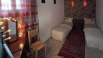 Ein kleiner, gemütlicher Raum mit einem Bett, einem Sessel, einem Schreibtisch mit Spiegel und Beleuchtung, einem roten Teppich und einer Steinwand mit Bildern.
