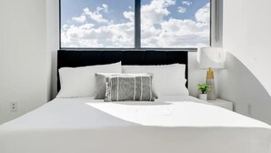 Una cama con ropa de cama blanca, una almohada a rayas, una mesita de noche con una lámpara y una planta pequeña.