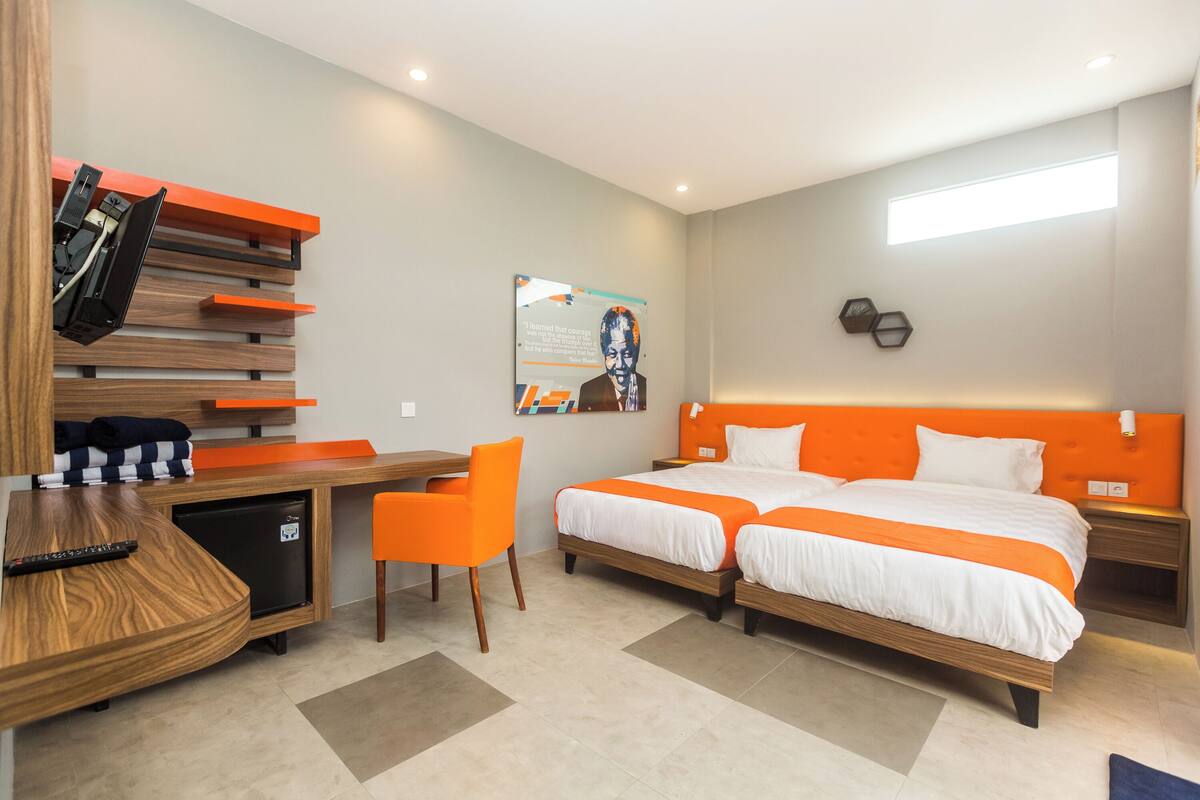 A hotel room with two beds, a desk, a chair, a small fridge, and a poster on the wall.