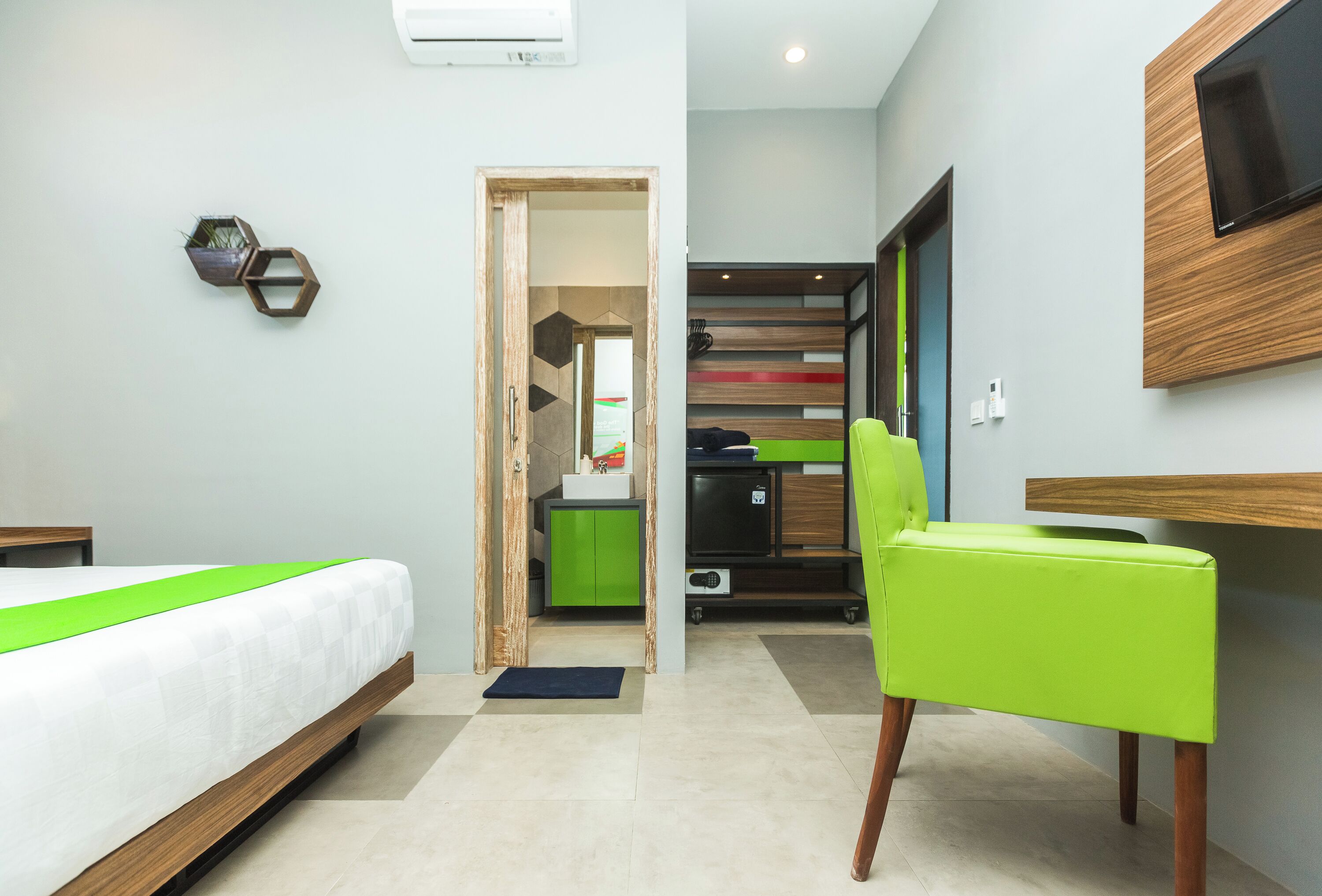 A modern hotel room with a bed, a green chair, a TV mounted on the wall, and a kitchenette in the background.