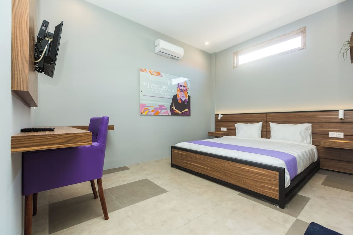 A hotel room with a bed, a desk with a chair, an air conditioner, and a poster on the wall.