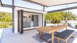 A modern outdoor patio with a wooden table and bench, a sliding glass door leading to a kitchen, and a view of greenery. - Little Cove Luxe - Naousa 10 (Noosa)