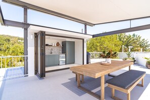 Une terrasse extérieure moderne, avec une table et un banc en bois, une porte-fenêtre coulissante donnant sur la cuisine et une vue sur la verdure.