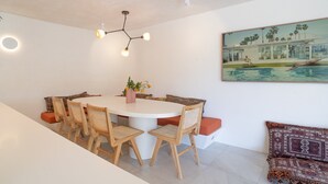 A dining area with a round table, wooden chairs, a modern chandelier, and a painting of a swimming pool. - Little Cove Luxe - Naousa 10 (Noosa)