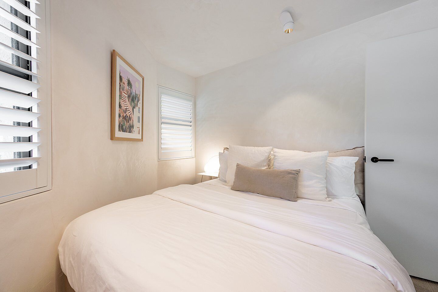 A bedroom with a large bed, white bedding, a single window with blinds, a wall-mounted light fixture, and a framed picture.