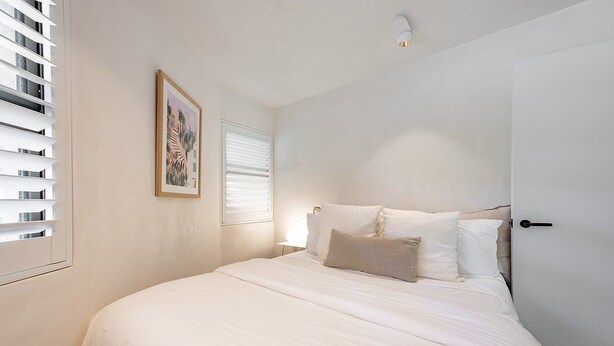 A bedroom with a large bed, white bedding, a single window with blinds, a wall-mounted light fixture, and a framed picture. - Little Cove Luxe - Naousa 10 (Noosa)