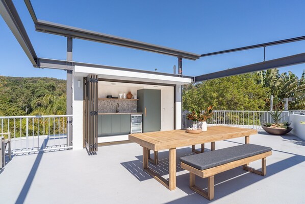 An outdoor patio with a wooden table and bench, a sliding glass door leading to a kitchen, and a view of a green landscape.