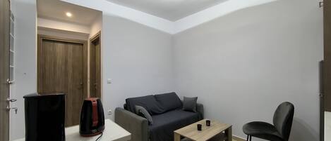 A modern living room with a sofa, a coffee table, and a chair. There is a TV on a stand and a door in the background.