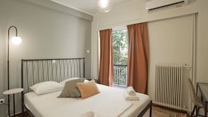Une chambre à coucher comprenant un lit, une table de chevet, une lampe, une fenêtre avec des rideaux, un radiateur et un appareil de climatisation.