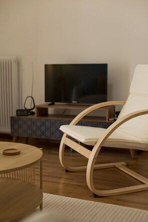 Ein modernes Wohnzimmer mit Holzboden, einem runden Holztisch, einem weißen, gepolsterten Sessel, einem Fernseher auf einem Holzschrank und einer Schrankwand mit Muster.