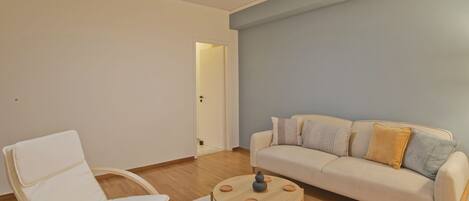 Una sala de estar con un sofá blanco, una silla blanca, una mesa de centro redonda de madera y una puerta que da a otra habitación.