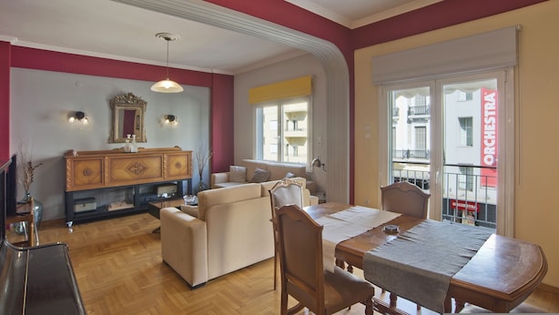 A dining area with a wooden table and chairs, a sofa, and a mirror above a wooden cabinet. - Baroness Apartment in Piraeus (Piraeus)